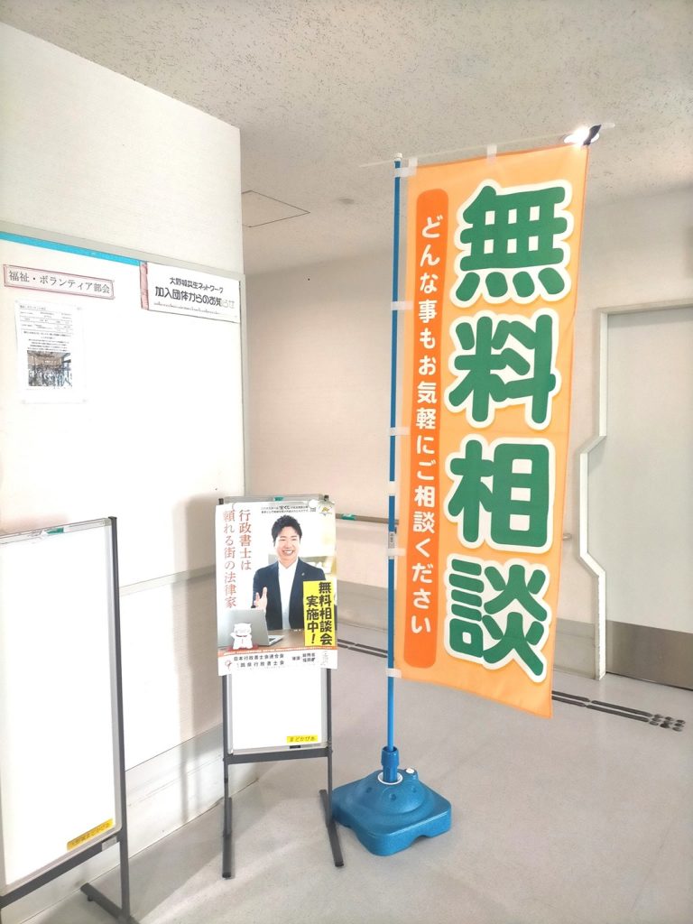 大野城まどかぴあ館内に設置された、行政書士による無料相談会のオレンジ色ののぼり旗と、男性相談員の写真が載ったイーゼルポスターの様子。